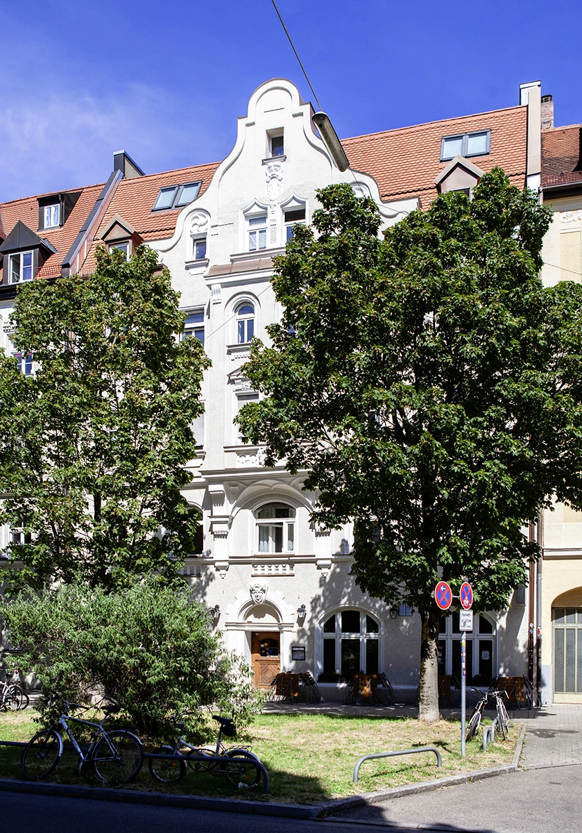 stoiber-architektur-muenchen-2026-sanierung-donnersbergerstr-02 Weiße Jugendstilfassade in München-Neuhausen mit Ziergiebel, Rundbogenportal und Bäumen im Vordergrund