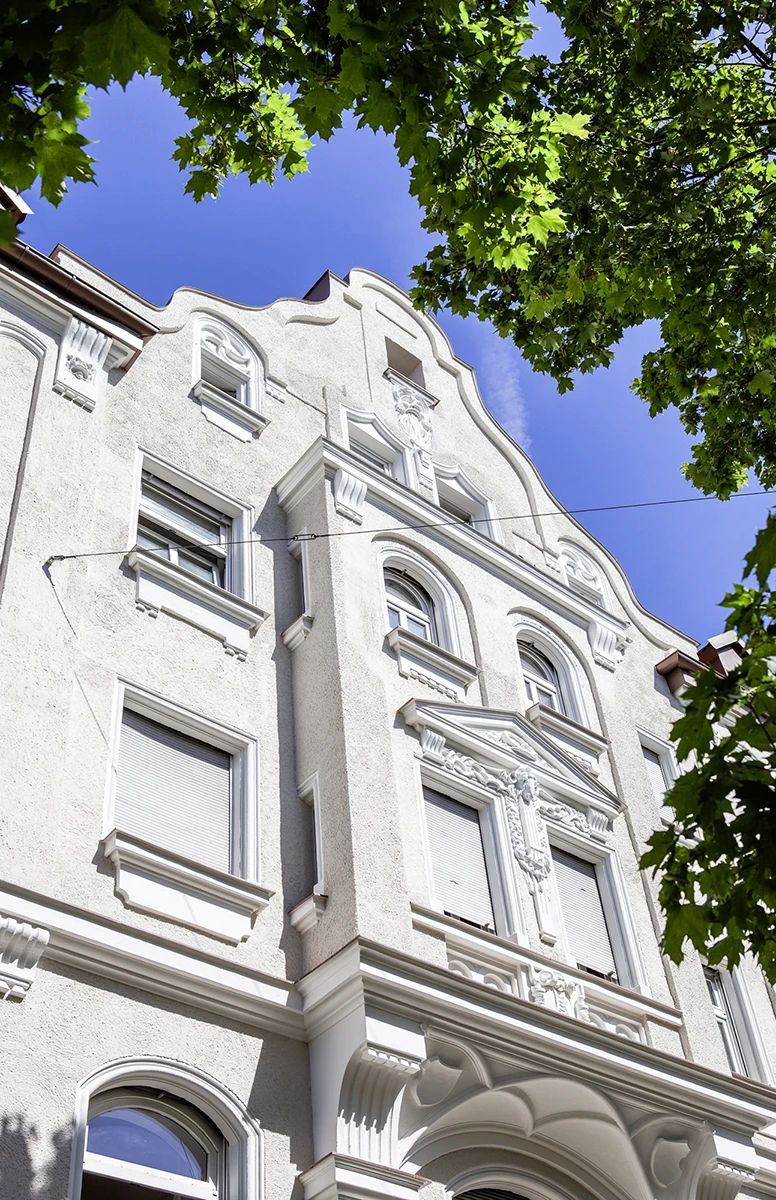 stoiber-architektur-muenchen-2026-sanierung-donnersbergerstr-03 Untersicht der weißen Stuckfassade mit Fensterverdachungen, Konsolen und geschwungenem Giebel unter Baumkronen