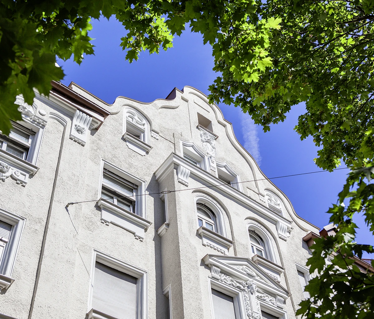 stoiber-architektur-muenchen-2026-sanierung-donnersbergerstr-04 Stuckverzierter Jugendstilgiebel mit Erker und Dreiecksgiebel-Fenster, eingerahmt von grünem Laub vor blauem Himmel