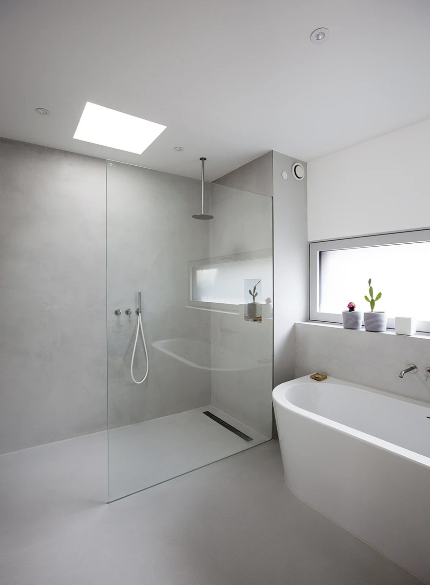 stoiber-architektur-neubau-einfamilienhaus_muenchen-badezimmer_13 Modernes Badezimmer in hellem Grau gefliest, Wände und Decke in Weiss, Lichtschacht über der bodenebenen Dusche, stylische Badewanne unter dem Fenster