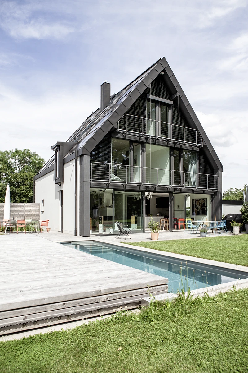 Blick auf das Haus vom Garten, komplette Fensterfront auf der Giebelseite, graues Dach, im Vordergrund ein halb zugedeckter Pool mit Holzumrandung