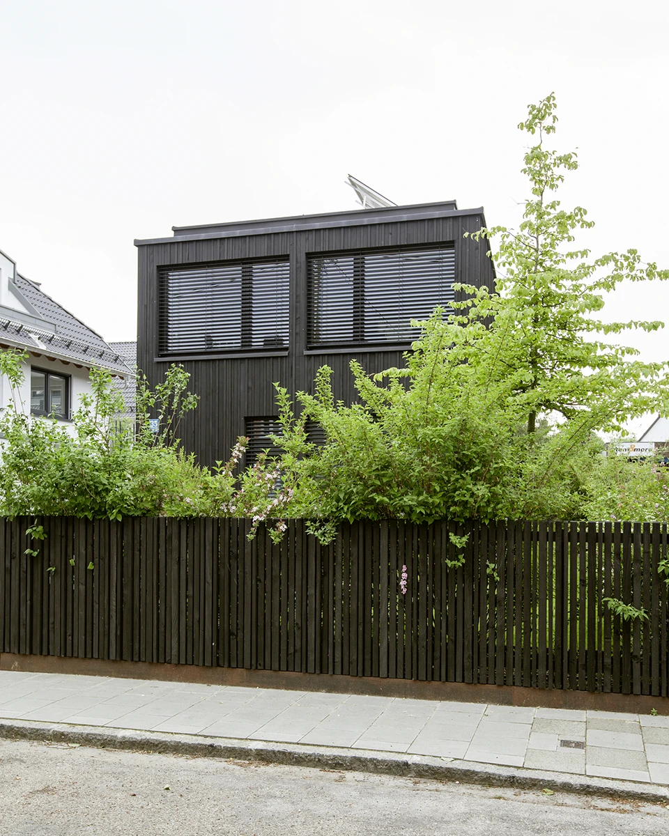 stoiber-architektur-neubau-einfamilienhaus_muenchen_aussen_03 Blick auf ein modernes Einfamilienhaus von der Strasse, Fassade mit dunklem Holz, große Fenster, Büsche und Bäume