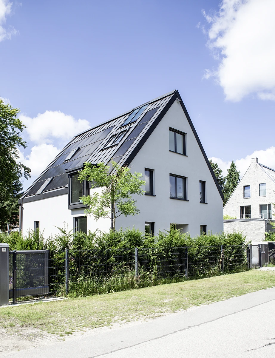 Blick auf das Haus von der Strasse, weisse Wände, graues Dach, Im Vordergrund eine Hecke hinter Doppelstabmattenzaun
