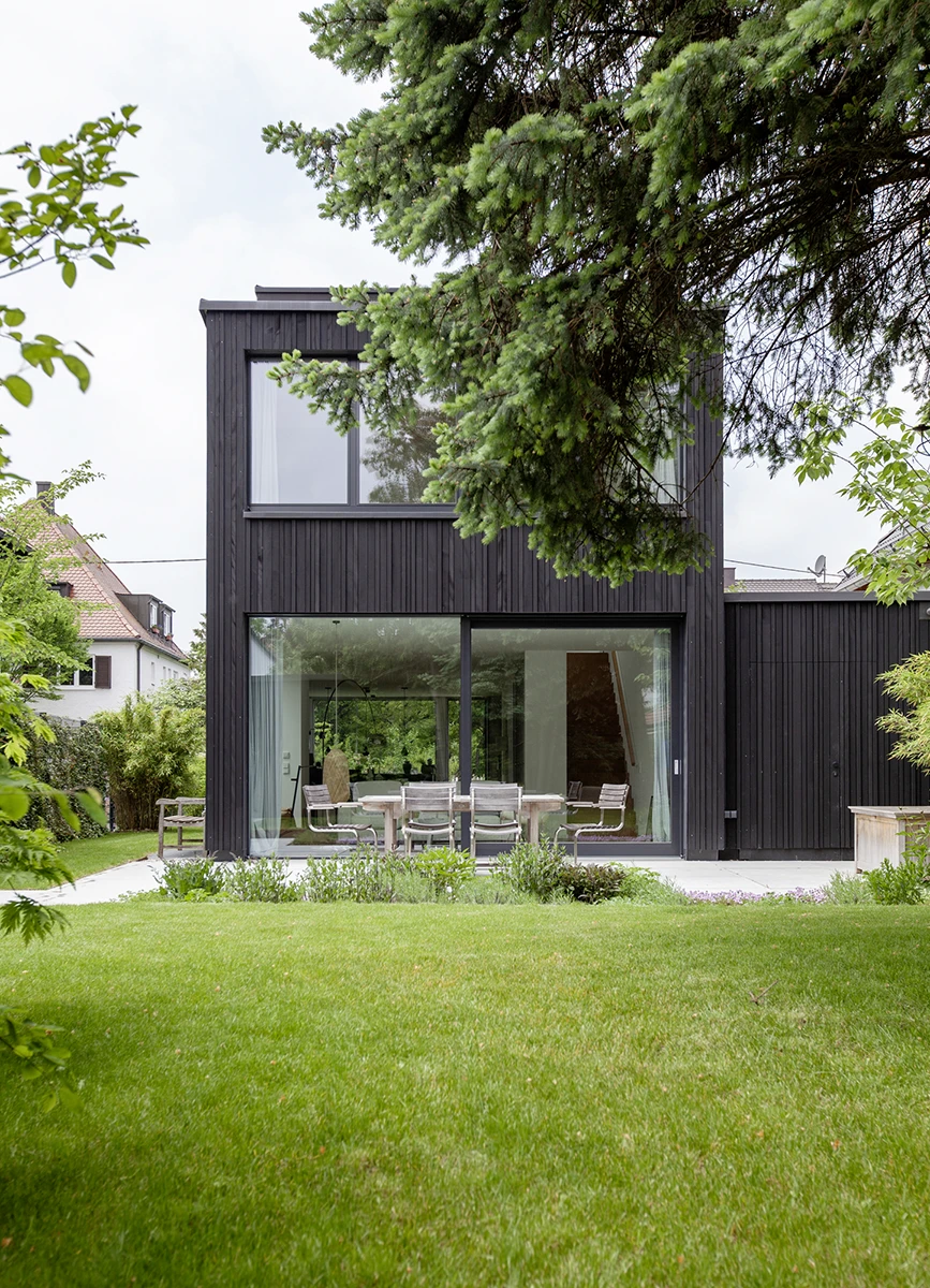 Blick auf ein modernes Einfamilienhaus vom Garten aus, Fassade mit dunklem Holz, große Fenster, Büsche und Bäume