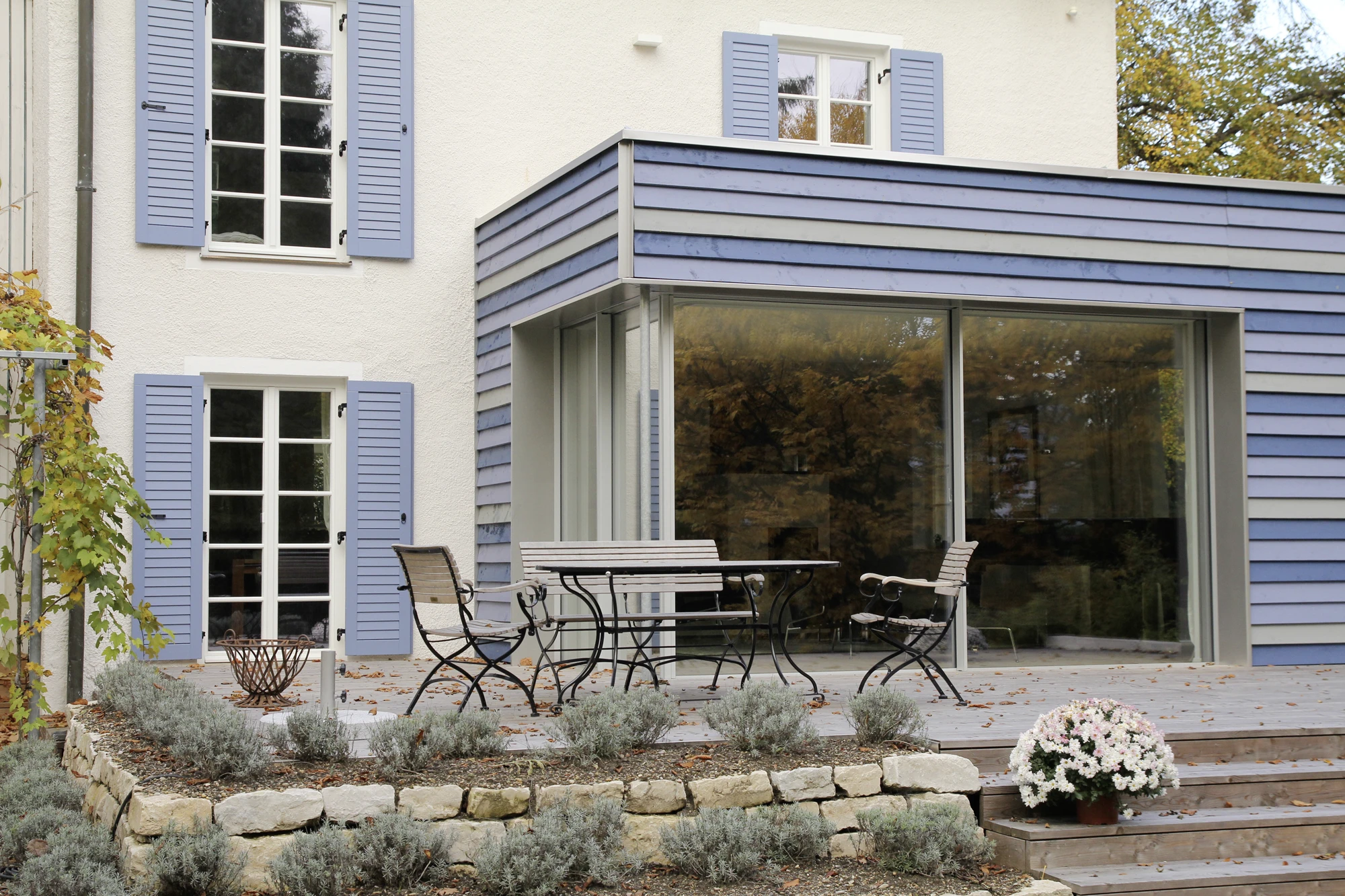 stoiber-architektur_sanierung-einfamilienhaus-anbau_muenchen-obermenzing_anbau-terasse_03 Blick auf den neuen Anbau aus dem Garten, Fassade des Anbaus in leicht bläulicher Holzverkleidung, Fassade des Hauses weiß verputzt
