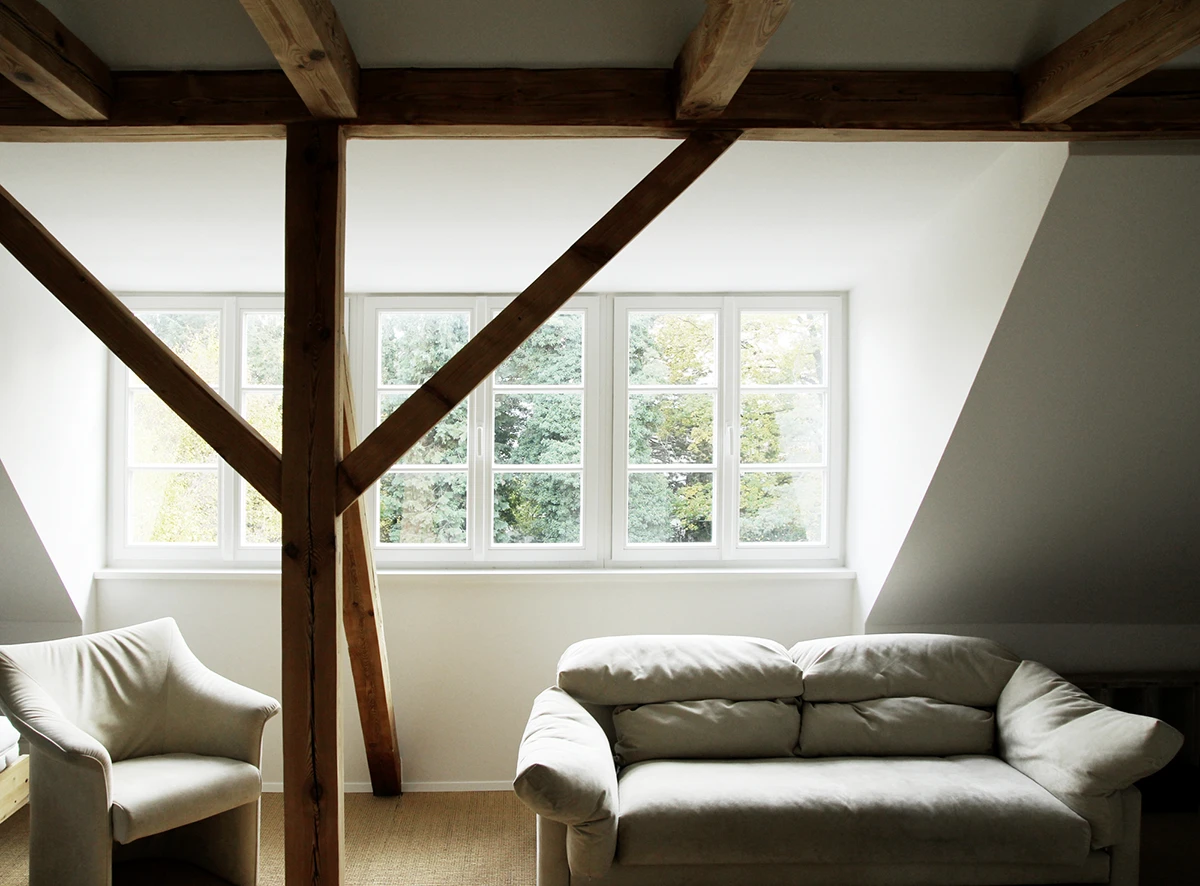 stoiber-architektur_sanierung-einfamilienhaus-anbau_muenchen-obermenzing_dachgeschoss_07 Dachgeschoss mit Sessel und Sofa, dunkle Sichtbalken und großem Fenster