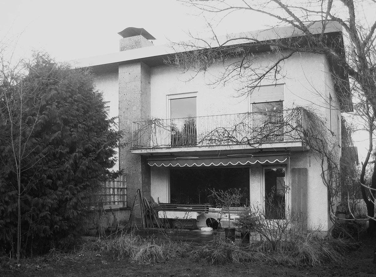 stoiber-architektur_sanierung-einfamilienhaus_muenchen_bestand_07 Blick auf das Haus vom Garten, vor der Sanierung, alter Putz, Balkon im ersten Stock, klassische Aufteilung im Erdgeschoss mit großem Fenster und kleiner Tür zur Terasse, Markiese