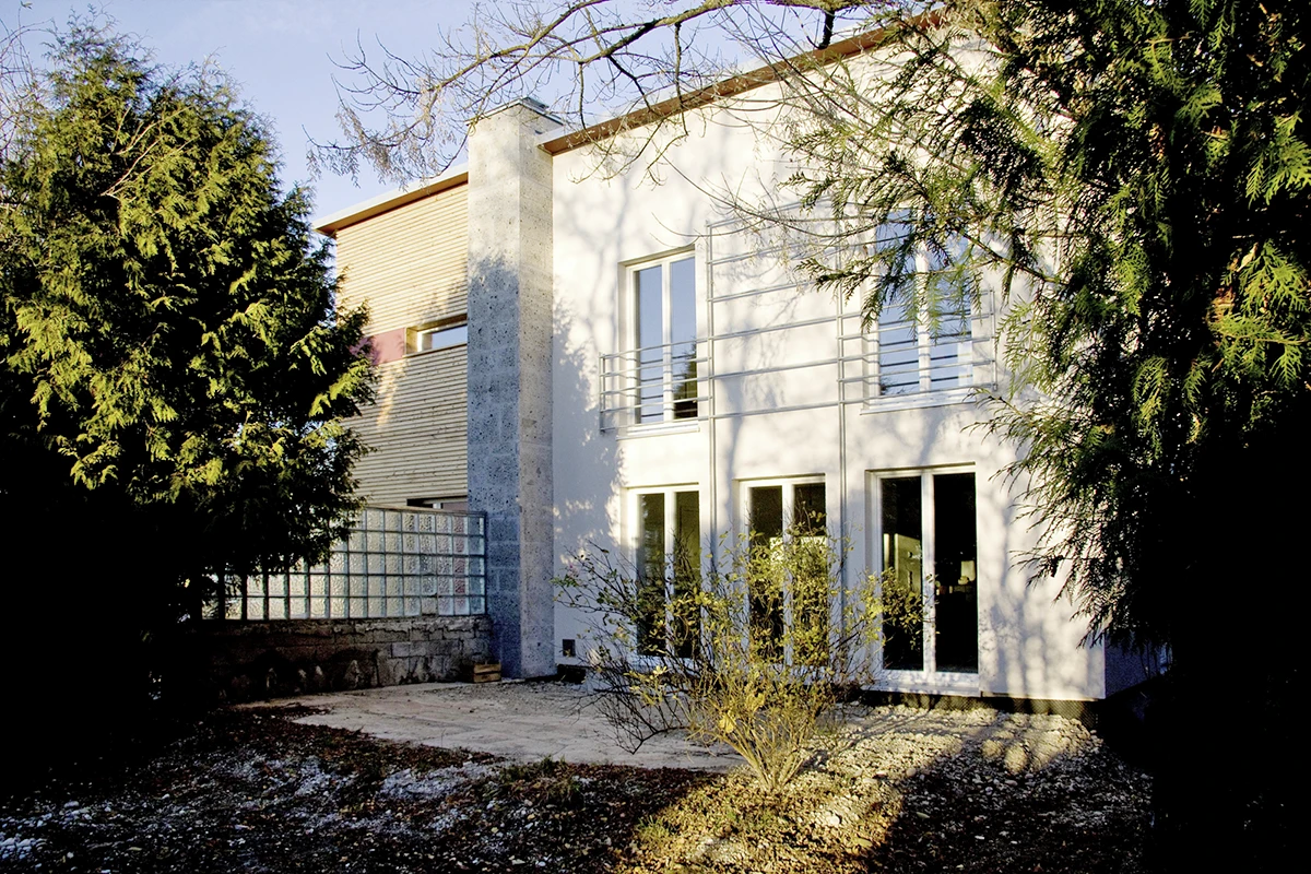 stoiber-architektur_sanierung-einfamilienhaus_muenchen_fassade-hinten_02 Blick auf das sanierte Einfamilienhaus von der Gartenseite, drei Türen im Erdgeschoss, zwei hohe Fenster im ersten Stock, Bäume links und rechts