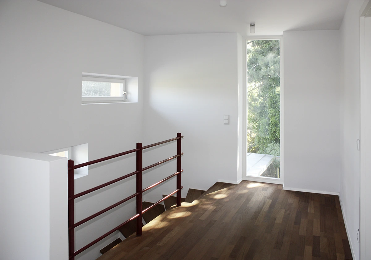 stoiber-architektur_sanierung-einfamilienhaus_muenchen_flur-treppenhaus_05 Lichtdurchfluteter Flur im ersten Stock, dunkler Holzfussboden, Treppenabgang links, am Ende des Ganges schmales deckenhohes Fenster