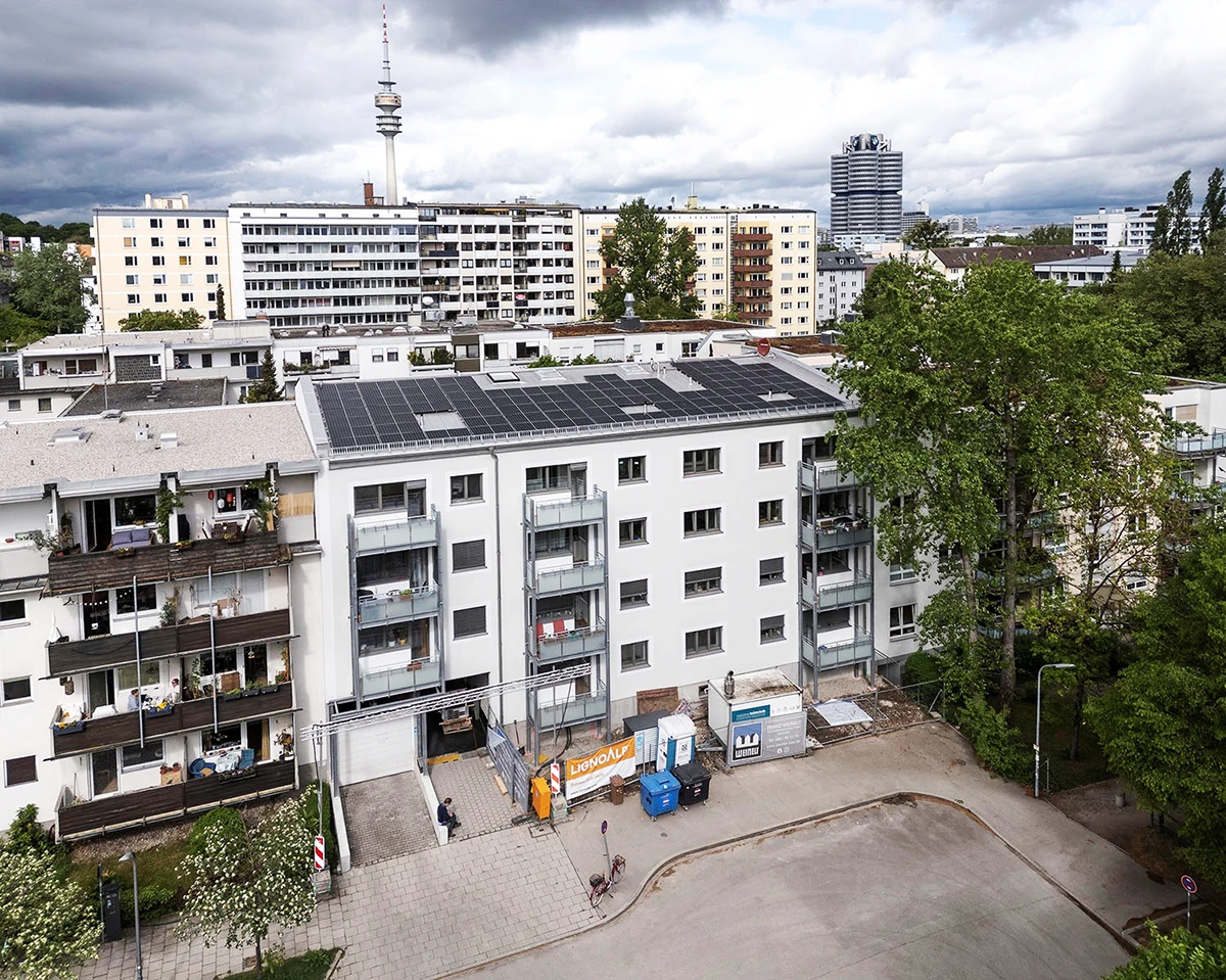 stoiber-architektur-muenchen-2026-sanierung-brunnerstr-baustart-03 Luftbild des sanierten Wohnhauses mit Photovoltaik auf dem Satteldach und neuen Stahlbalkonen, dahinter der Olympiaturm