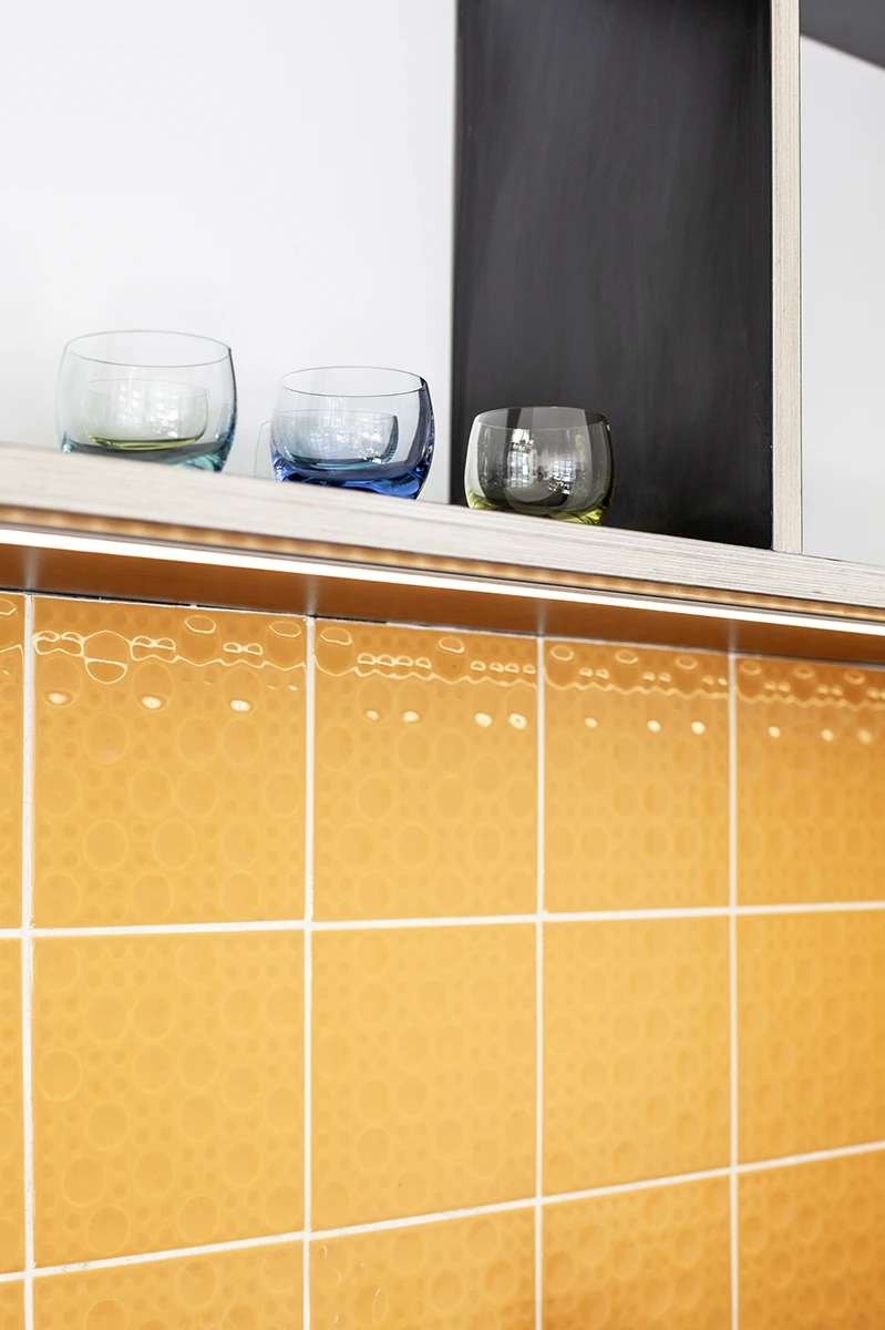 stoiber-architektur-muenchen-2026-sanierung-gudrunstr-01 Orangefarbene Reliefkacheln mit Kreismuster unter einem Holzregal mit farbigen Trinkgläsern