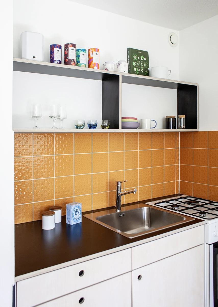 stoiber-architektur-muenchen-2026-sanierung-gudrunstr-03 Küchenzeile mit Edelstahlspüle und Gasherd vor orangefarbenen Fliesen, darüber ein offenes Regal mit Teedosen und Geschirr