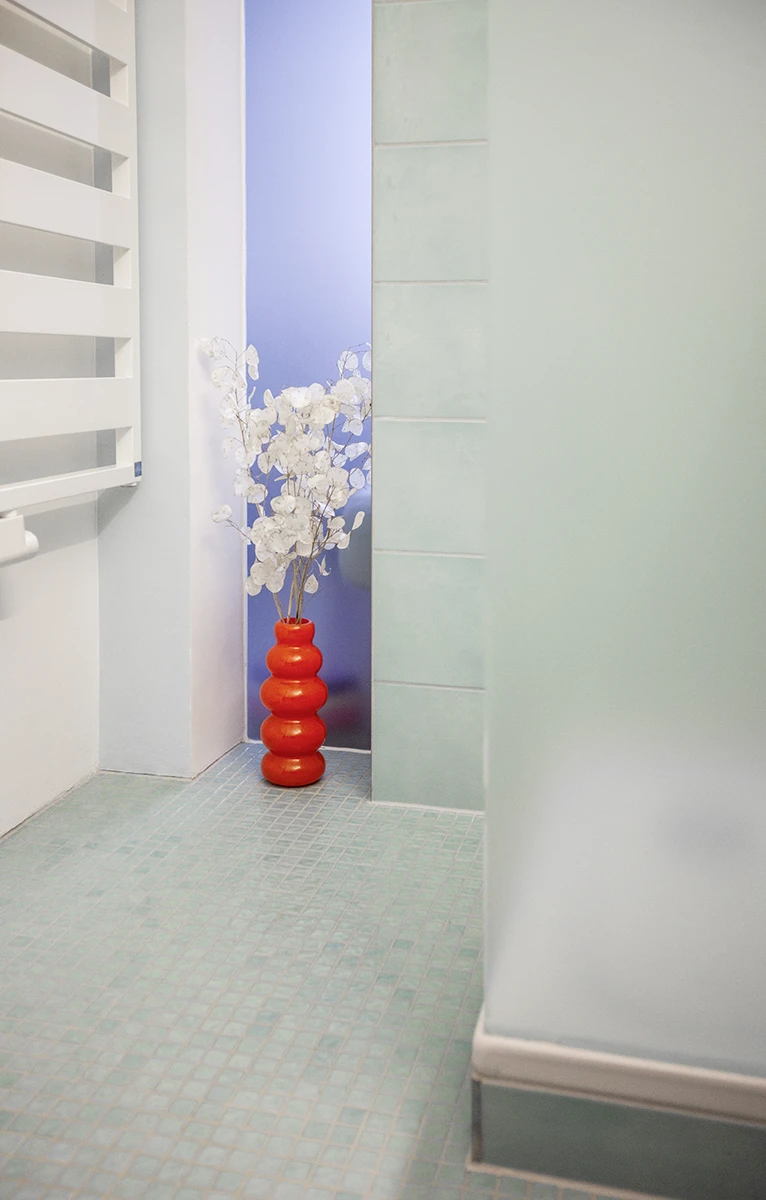 stoiber-architektur-muenchen-2026-sanierung-gudrunstr-09 Schmaler Lichtschlitz mit satiniertem Glas in der Badwand, davor eine rote Vase mit weißen Zweigen auf Mosaikboden
