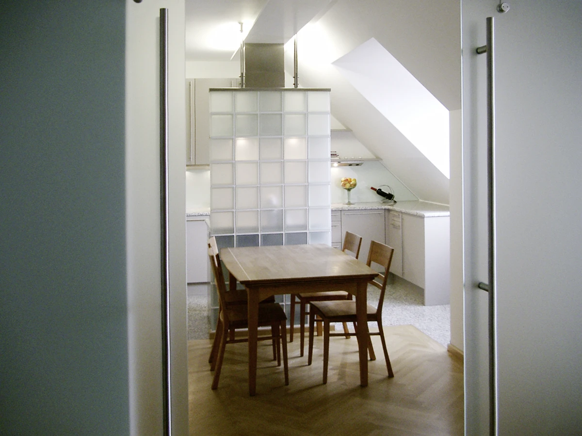 stoiber-architektur_denkmal_sanierung_dachgeschossausbau_muenchen_kueche-essbereich_05 Holzesstisch mit vier Stühlen vor einer Glasbausteinwand, dahinter eine helle Küche unter der Dachschräge