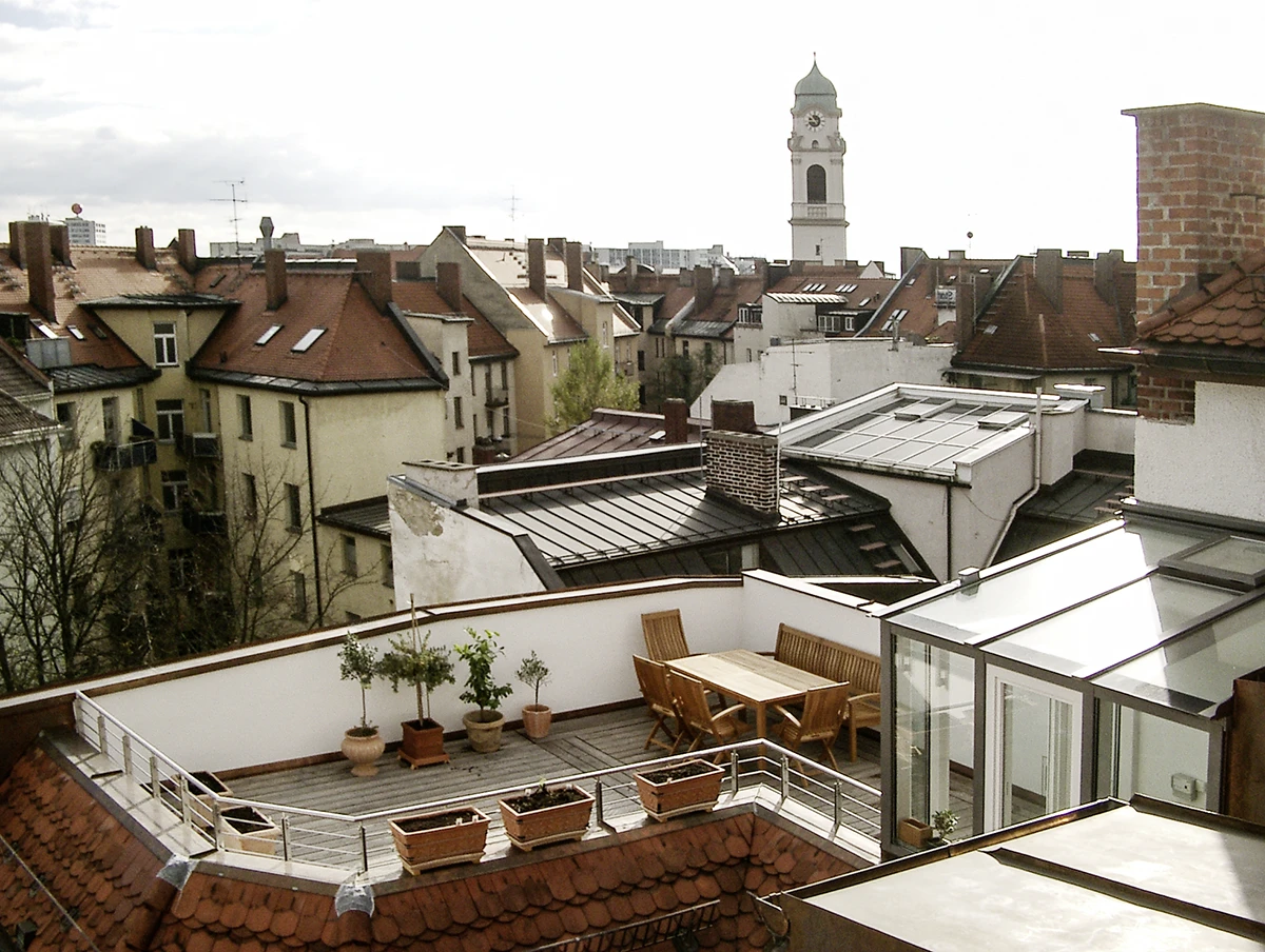 stoiber-architektur_denkmal_sanierung_dachgeschossausbau_muenchen_von oben_01 Dachterrasse mit Holzmöbeln und Pflanzkübeln über den Dächern von München-Haidhausen, im Hintergrund ein Kirchturm